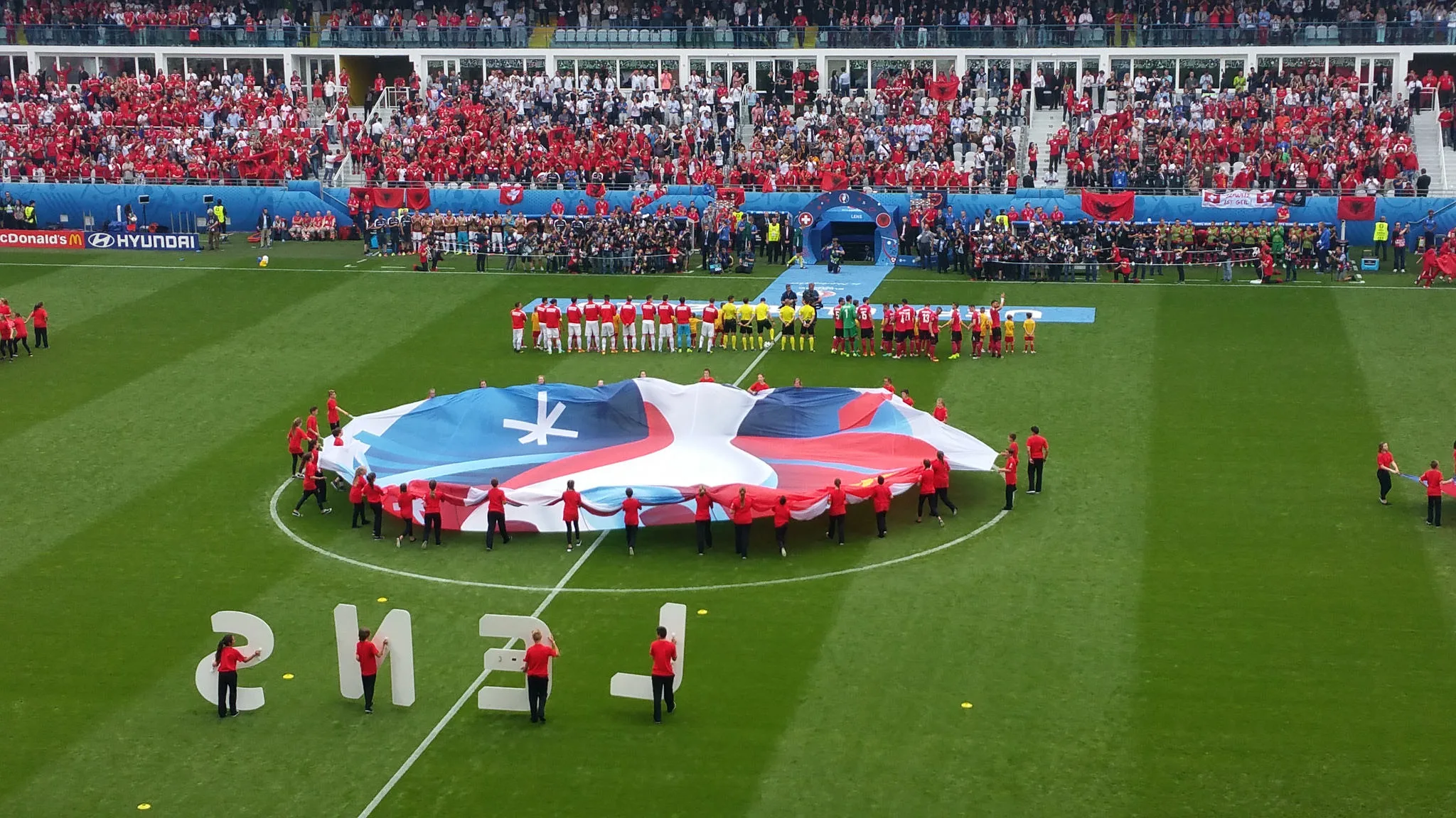 Cerimonia prima del calcio d’inizio a una partita degli Europei 2016 a Lens: bandiera gigante nel cerchio di centrocampo, le squadre schierate