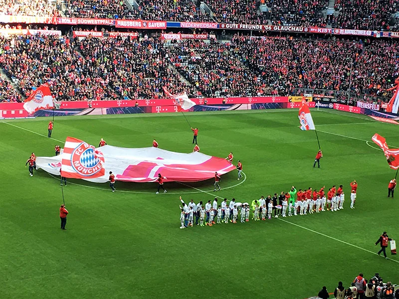 Bandiera gigante con lo stemma del FC Bayern sul prato dell’Allianz Arena, le squadre schierate prima del calcio d’inizio