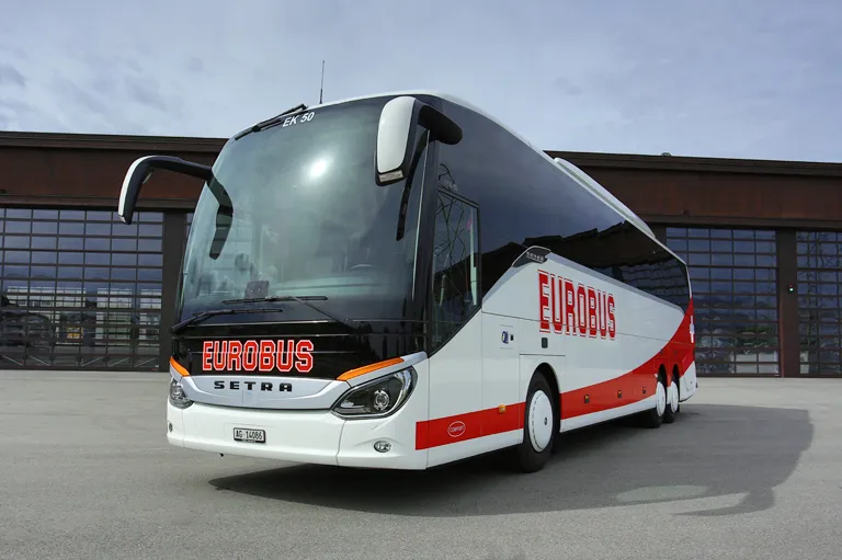 A white EUROBUS Comfort coach with a red wave parked in front of a hall