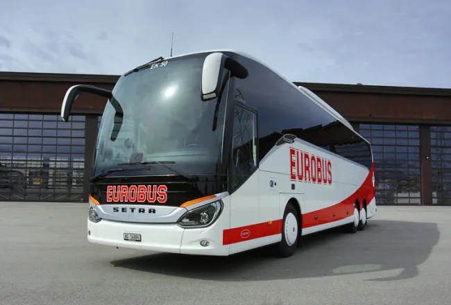 A white EUROBUS Comfort coach with a red wave parked in front of a hall