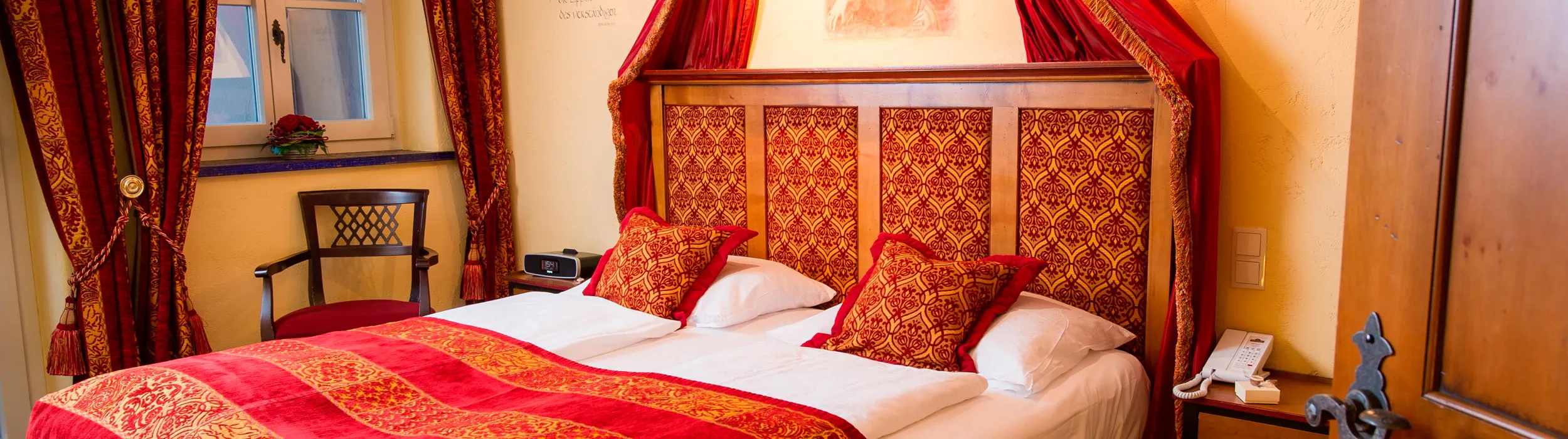 Un lit double avec des coussins et un jeté rouge et or sous un baldaquin rouge, à côté d’une fenêtre aux lourds rideaux