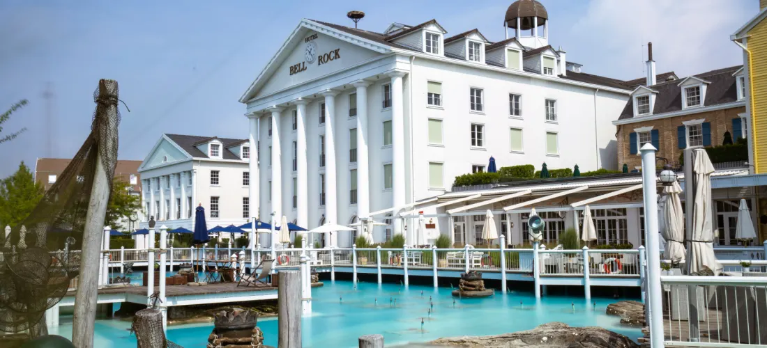 Il bianco Hotel Bell Rock con portale a colonne dietro una vasca d’acqua turchese con passerelle di legno e ombrelloni
