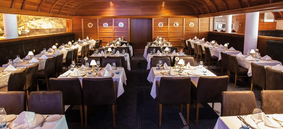 Un ristorante con tavoli apparecchiati di bianco, sedie imbottite marroni e pareti rivestite in legno con oblò