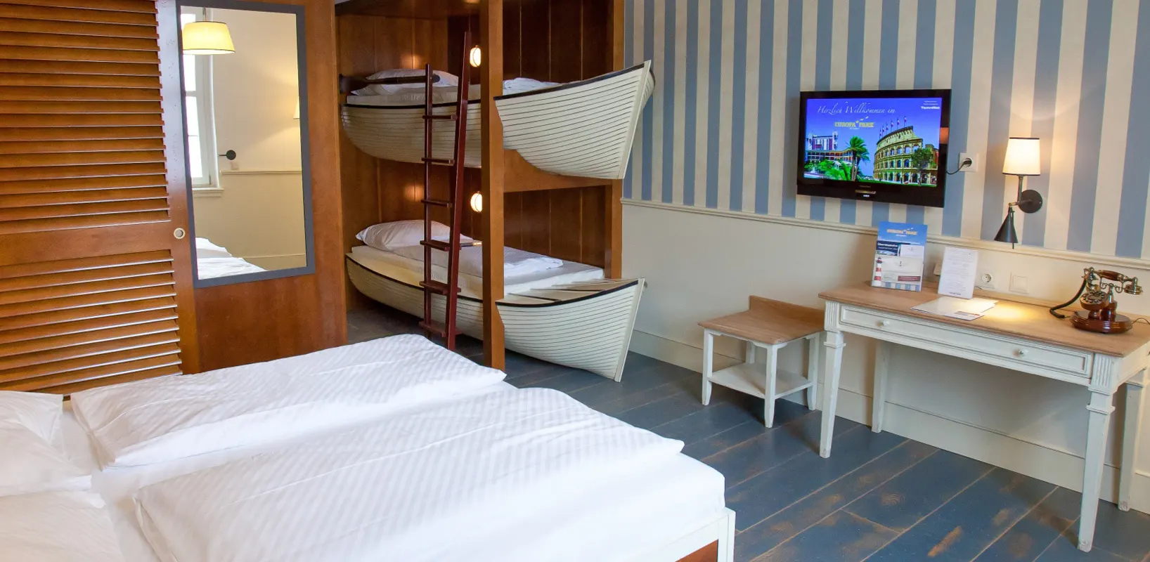 Una camera d’hotel con letto matrimoniale, due letti a forma di barche bianche uno sopra l’altro e carta da parati a righe blu e bianche