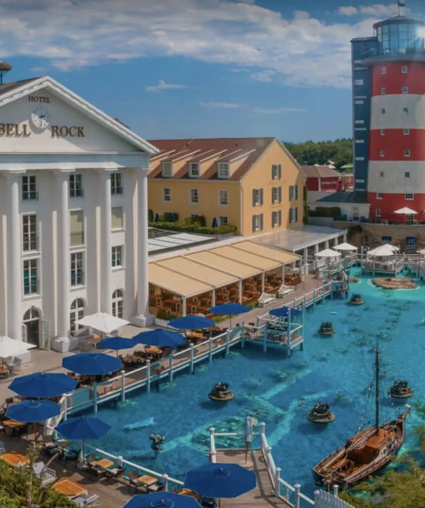 Il bianco Hotel Bell Rock con portale a colonne, accanto un faro bianco e rosso e una vasca d’acqua turchese con passerelle e una barca di legno