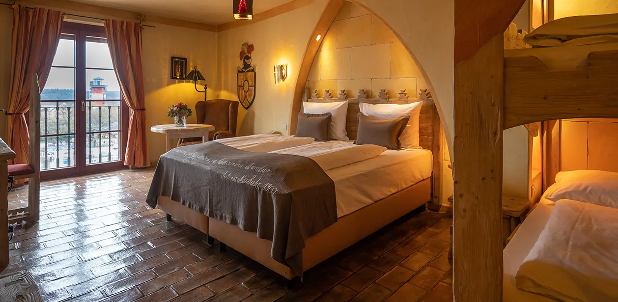 A hotel room with a double bed beneath a stone arch, a terracotta floor, a balcony door and a bunk bed in light wood