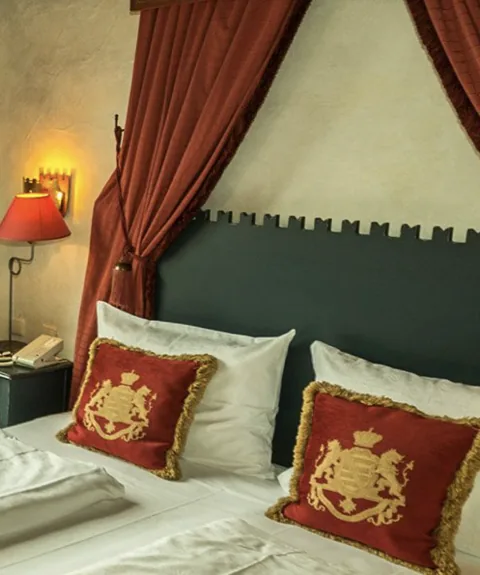 Un letto con cuscini rossi con stemma dorato sotto un baldacchino rosso, dietro una testiera scura con merli