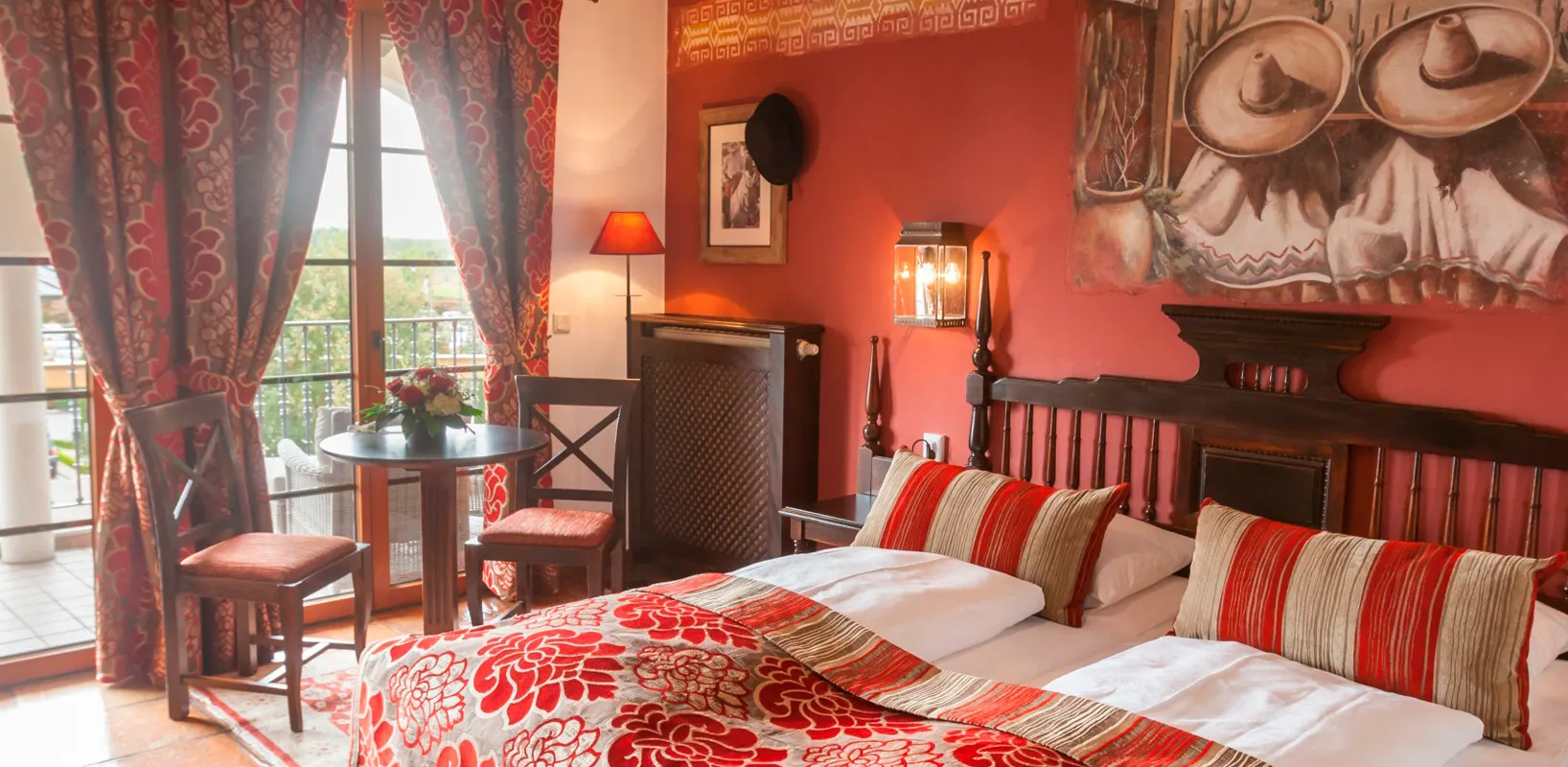 A hotel room with a red wall, a double bed with striped cushions and a mural with sombreros, on the left a balcony door with a table and two chairs
