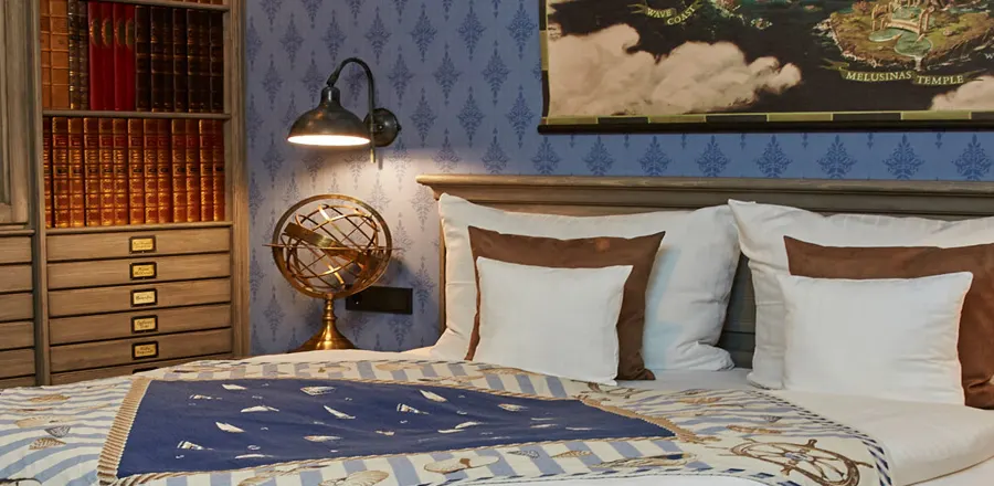Una camera d’hotel con letto matrimoniale sotto una vecchia carta geografica, carta da parati blu con motivi, un mappamondo e una cassettiera con libri