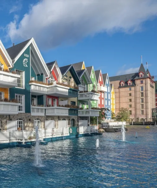 Casette di legno colorate con balconi su una vasca d’acqua con fontane, dietro un edificio in mattoni con torrette