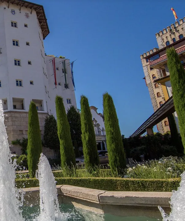 Un edificio bianco con bandiere dietro cipressi e una fontana, a destra una torre di castello con merli